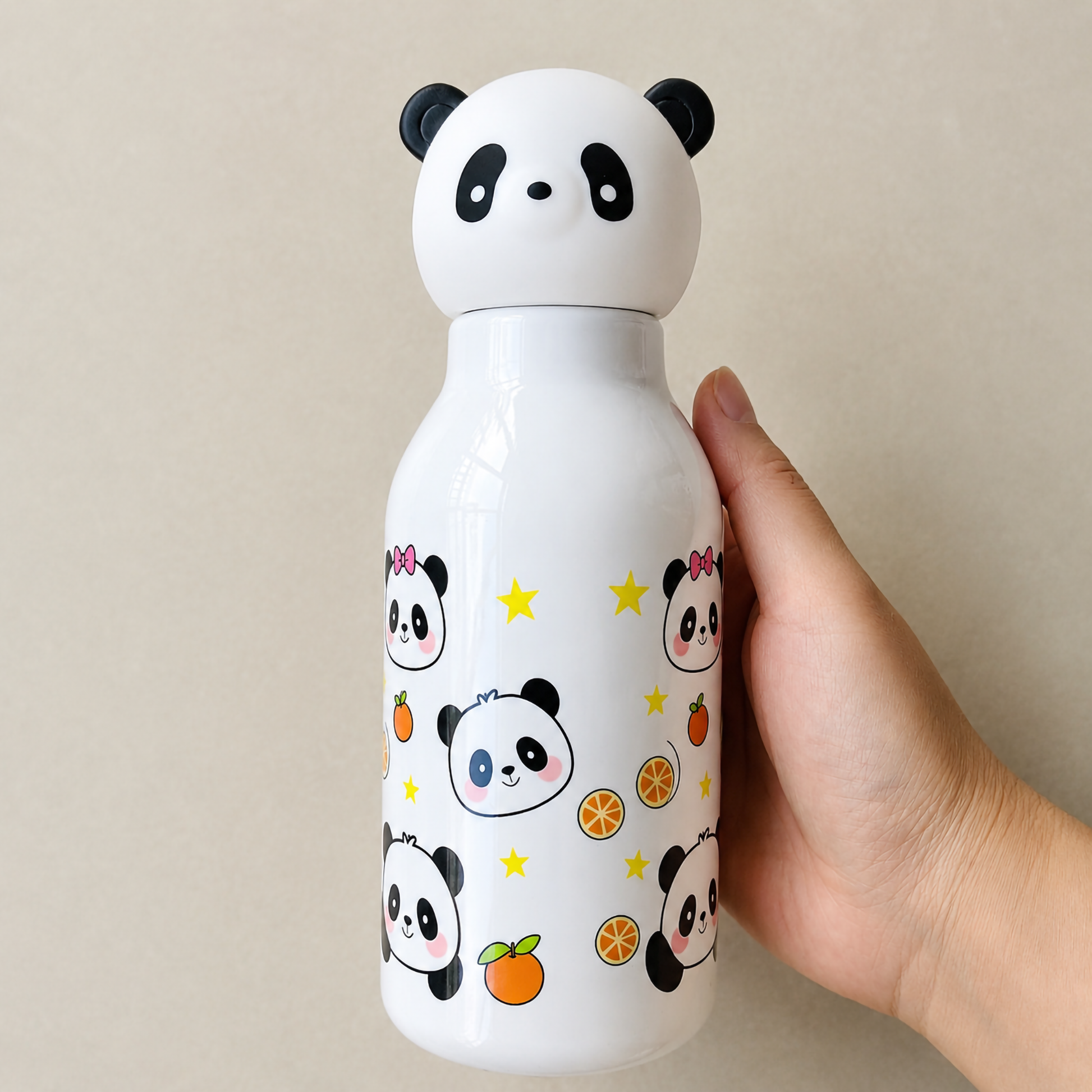 A hand holding a crisp white 500ml kids plastic water bottle covered in cute cartoon panda faces, yellow stars, and orange slices under a 3D panda cap