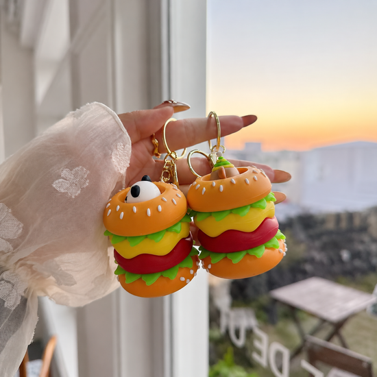 A close-up shot of a hand holding two silicone hamburger mini pouches with bunny and bear characters, showcasing the detailed 3D design against a warm sunset background.
