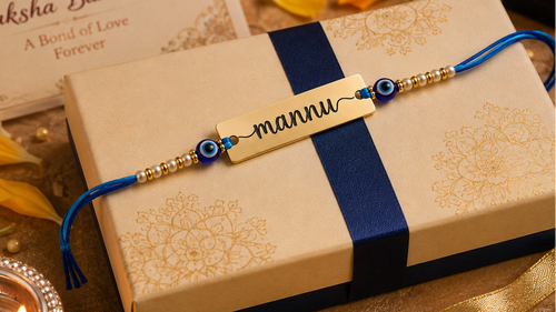 A personalized Rakhi with a gold nameplate engraved 'mannu' and blue evil eye beads, laid across a beige and navy gift box. The scene is decorated with a 'Happy Raksha Bandhan' card, orange marigold flowers, a silver diya, and other gift boxes.
