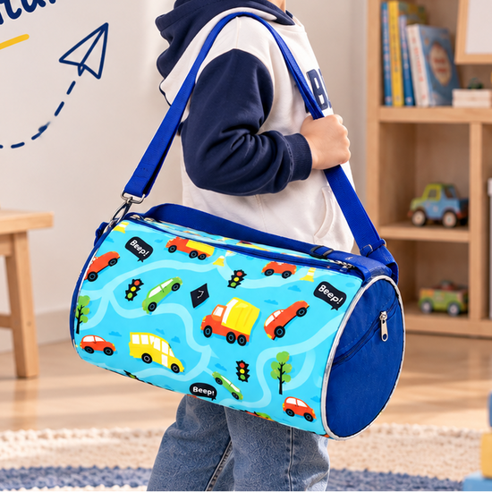 A young boy wearing a navy and white hoodie, jeans, and sneakers is shown in a bright children's room, carrying a light blue cylindrical duffel bag with a multicolor road-pattern print and blue straps. The image includes the text 'Small Bag Big Adventures' and is decorated with toys, books, and a teddy bear.