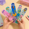 Five assorted liquid floating glitter ruler scales featuring pink rainbow, blue dog, space rocket, ocean octopus, and ice cream themes held up in two hands.