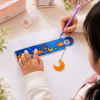 :A child using a blue space rocket-themed liquid quicksand scale with a hanging moon pendant to draw a straight line in a notebook