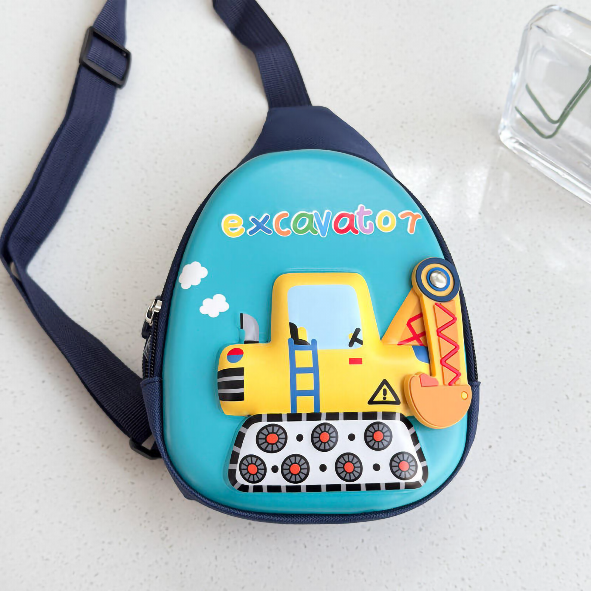 A close-up front view of a teal hardshell kids' crossbody bag showcasing a yellow excavator graphic with tread wheels