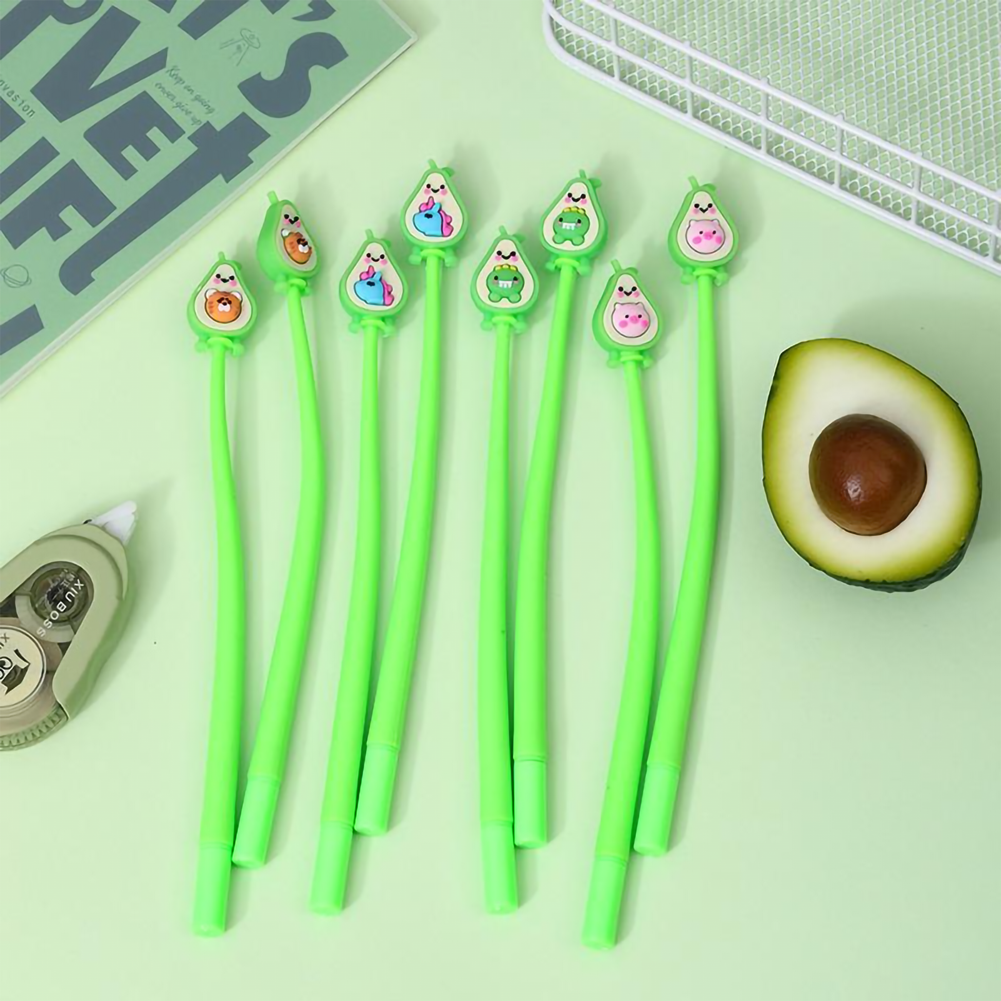 A collection of green barrel avocado shake gel pens displayed next to an open notebook and a fresh half-avocado on a light green background