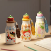 Three cute cartoon water bottles with fruit-themed caps and colorful handles are arranged on a wooden desk next to paintbrushes and an open sketchbook, with a lamp and blurred bookshelf in the background.