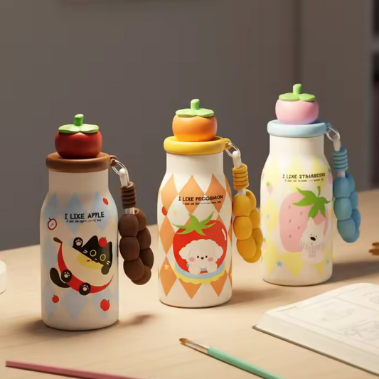 Three cute cartoon water bottles with fruit-themed caps and colorful handles are arranged on a wooden desk next to paintbrushes and an open sketchbook, with a lamp and blurred bookshelf in the background.