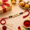 Flat lay of a personalized gold-finish name Rakhi with the name "mannu", blue thread, and evil eye beads arranged on a festive background with a diya, sweets, and a Happy Rakshabandhan card.