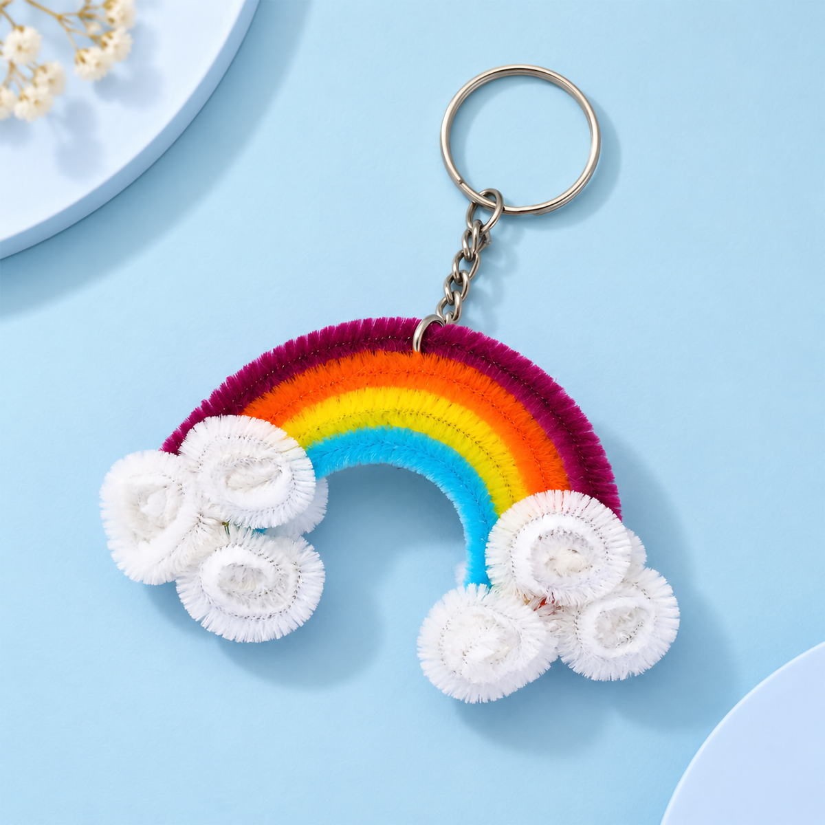 A handmade pipe cleaner rainbow keychain featuring multi-colored arch bands and fluffy white cloud bases laid flat on a light background.