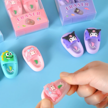 Multiple clear individual boxes containing pairs of cute 3D miniature clog slipper erasers in pink bunny, blue dinosaur, and purple character designs on a soft blue surface.