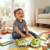 Toddler sitting on a colorful mat playing with a green interactive dinosaur piano toy.