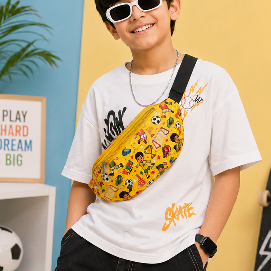 A boy wearing a white graphic t-shirt, black cargo pants, and white sunglasses models a yellow patterned crossbody fanny pack. The setting is a colorful studio with a yellow and blue wall, a neon lightning bolt sign, a skateboard, and a soccer ball.