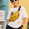 A boy wearing a white graphic t-shirt, black cargo pants, and white sunglasses models a yellow patterned crossbody fanny pack. The setting is a colorful studio with a yellow and blue wall, a neon lightning bolt sign, a skateboard, and a soccer ball.